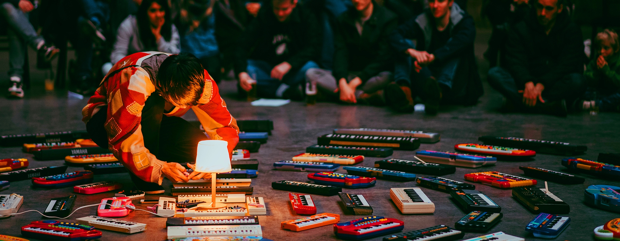 Artist crouching on floor surrounded by dozens of mini keyboards.